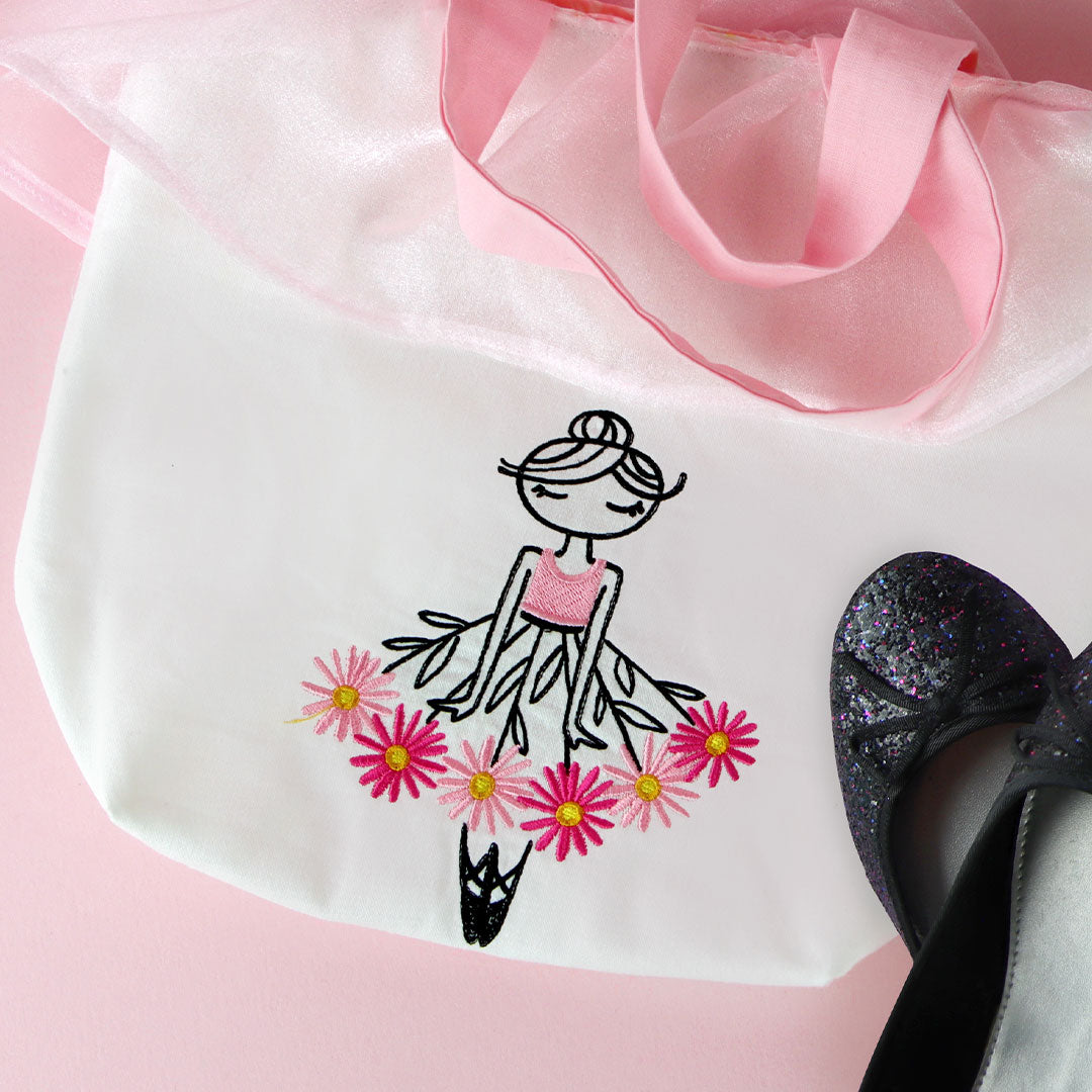 White bag featuring embroidered ballerinas in motion with floral accents, displayed on a pink background.