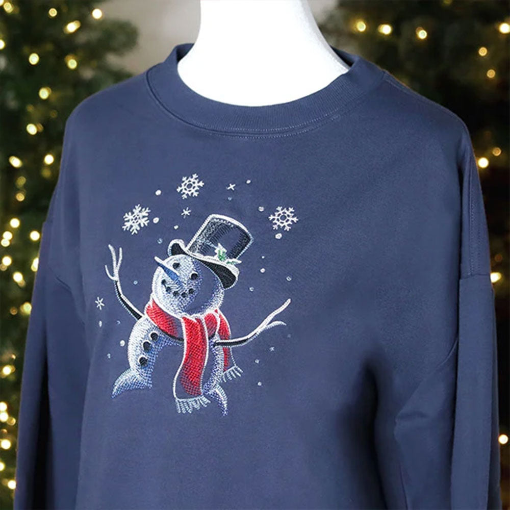 Digitally downloadable machine embroidery design of a cheerful snowman wearing a black top hat and red scarf, surrounded by falling snowflakes, on blue fabric.