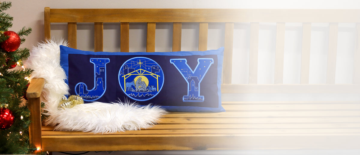 A blue rectangular pillow with the word "JOY" embroidered in light and dark blue and yellow, featuring a nativity scene inside the "O," rests on a wooden bench next to a white fur throw.
