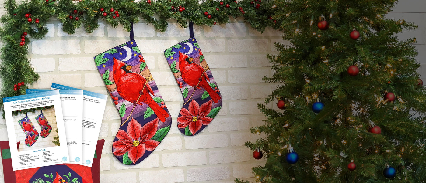 Two machine embroidered Christmas stockings with a vibrant red cardinal and red poinsettia pattern hanging on a white brick fireplace next to a decorated Christmas tree. A sewing pattern booklet is visible in the lower-left corner.
