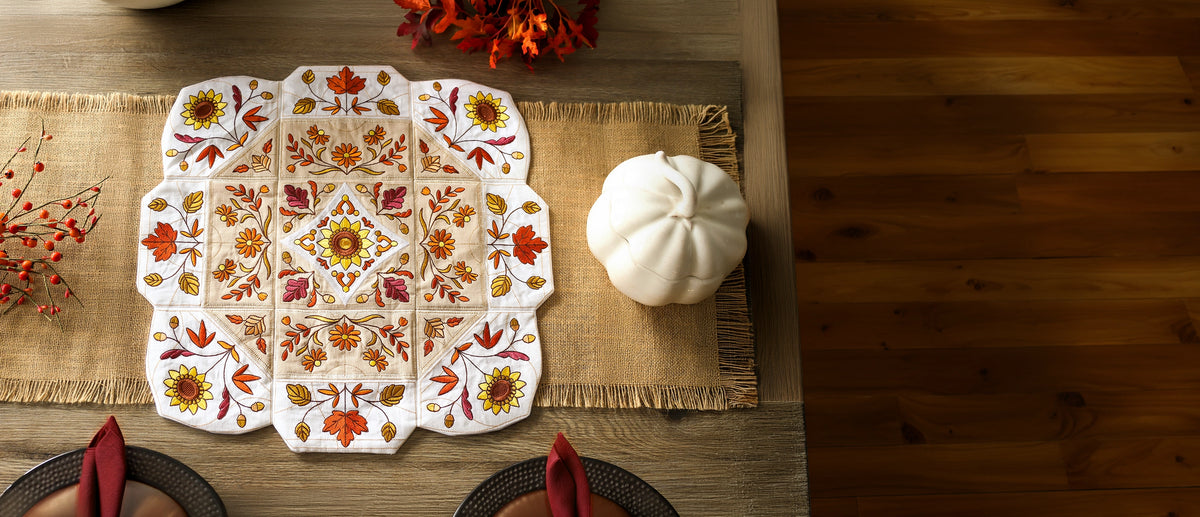 An autumn-themed machine embroidered table runner with a central medallion design, featuring orange, red, yellow, and green floral motifs on a white background. The runner is styled on a wooden table with a burlap placemat, a white pumpkin, and place settings, with a dark wood floor visible on the right.