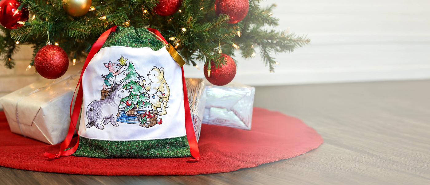 A Christmas-themed drawstring gift bag with a white front panel featuring a Pooh Baer & Friends illustration of a bear and a dog by a Christmas tree, with green patterned sides and red ribbons, placed under a decorated Christmas tree next to wrapped presents on a red tree skirt.