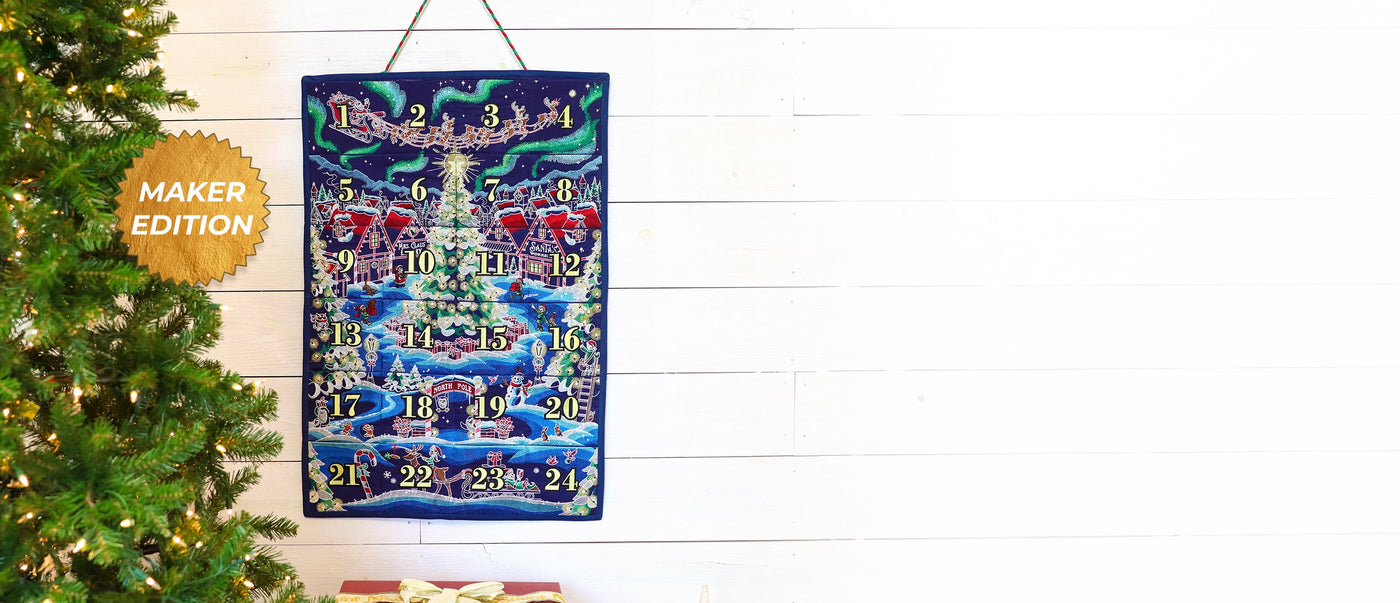 Machine embroidered advent calendar with Christmas scene design on a white wall next to a decorated Christmas tree.