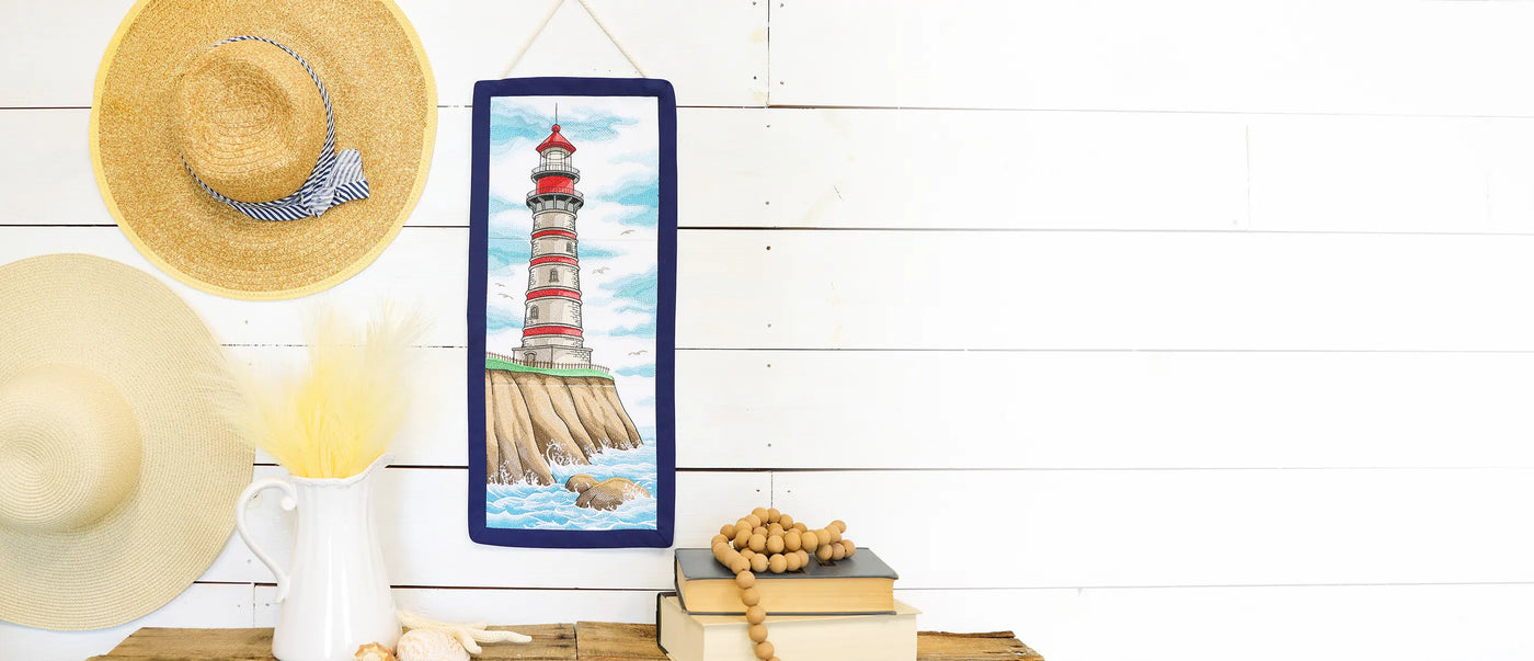Machine embroidered lighthouse wall hanging on a shelf with books and a hats against a white wooden background.