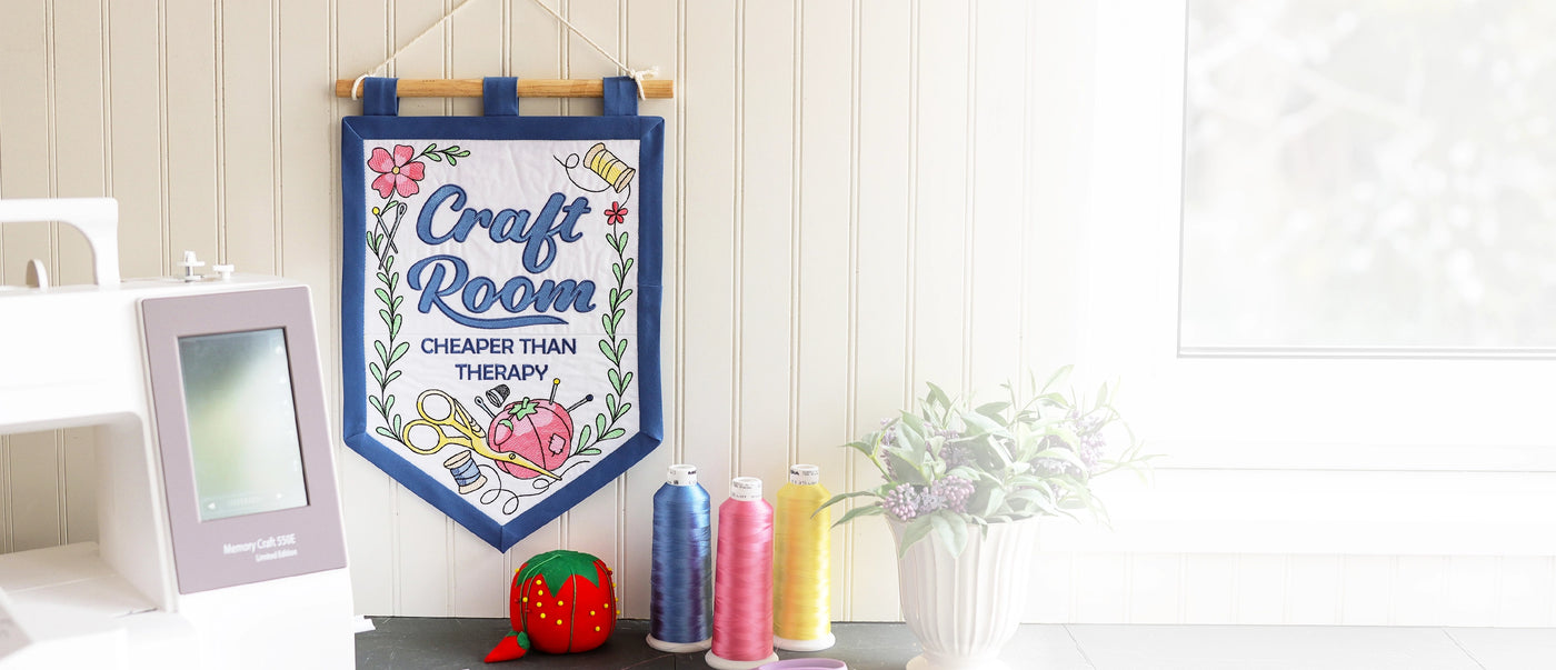 A decorative machine embroidered wall banner hanging on a white paneled wall in a craft room setting. The banner features a blue border and reads 'Craft Room CHEAPER THAN THERAPY' with colorful floral and sewing machine embroidery.