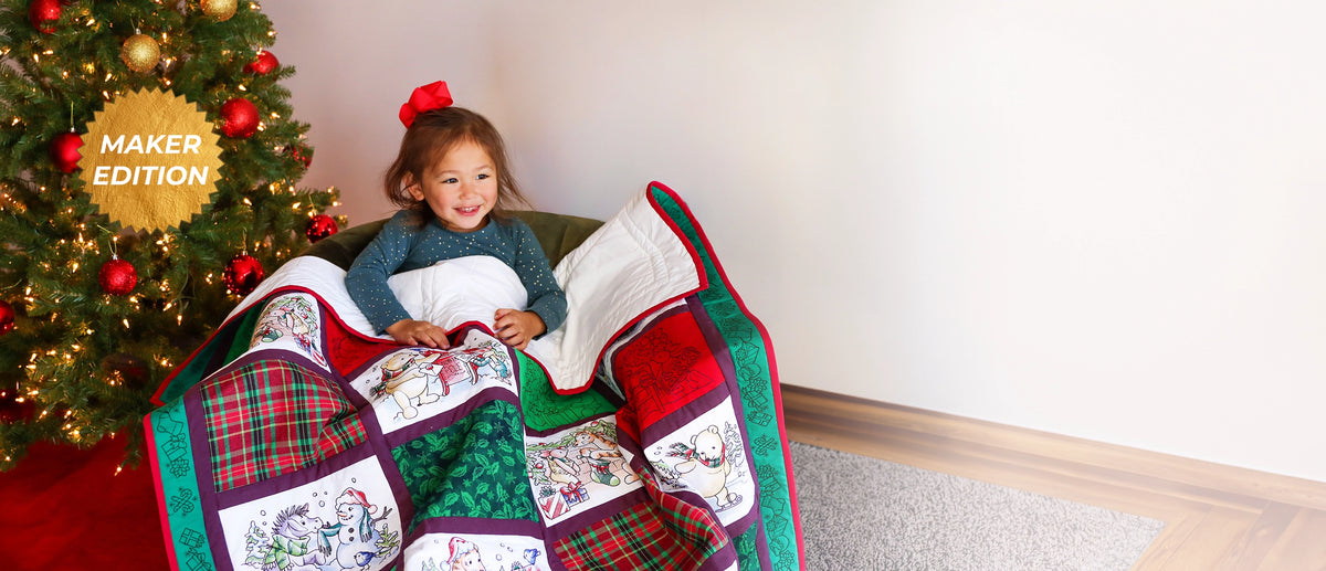 A child sits on a couch next to a decorated Christmas tree, covered with a handmade machine embroidered quilted blanket featuring pooh bearh & friends holiday patches and plaid patterns. A gold 'MAKER EDITION' badge appears on the tree, and the scene is set in a cozy living room.