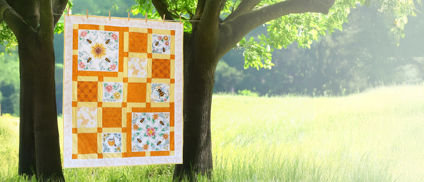Quilt with machine embroidery bee and floral designs hanging on a clothesline next to trees and a meadow.