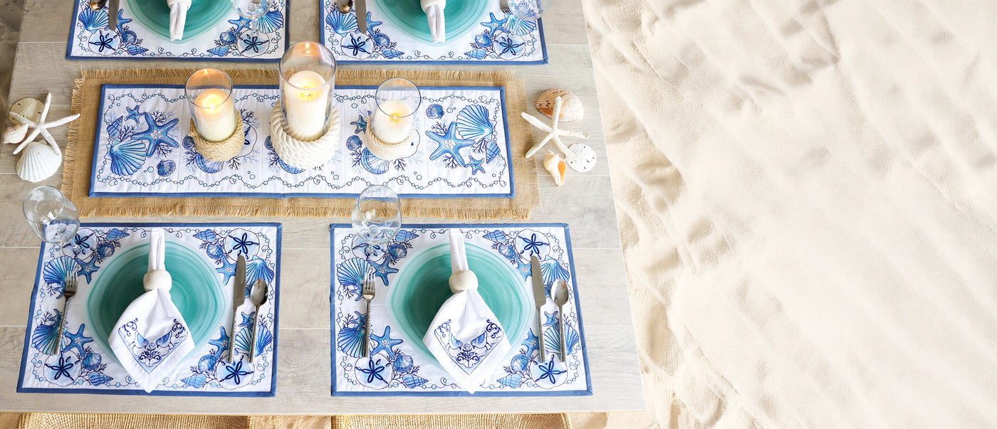 Table setting with blue and white machine embroidered place mats, candles, and cutlery on a textured surface.