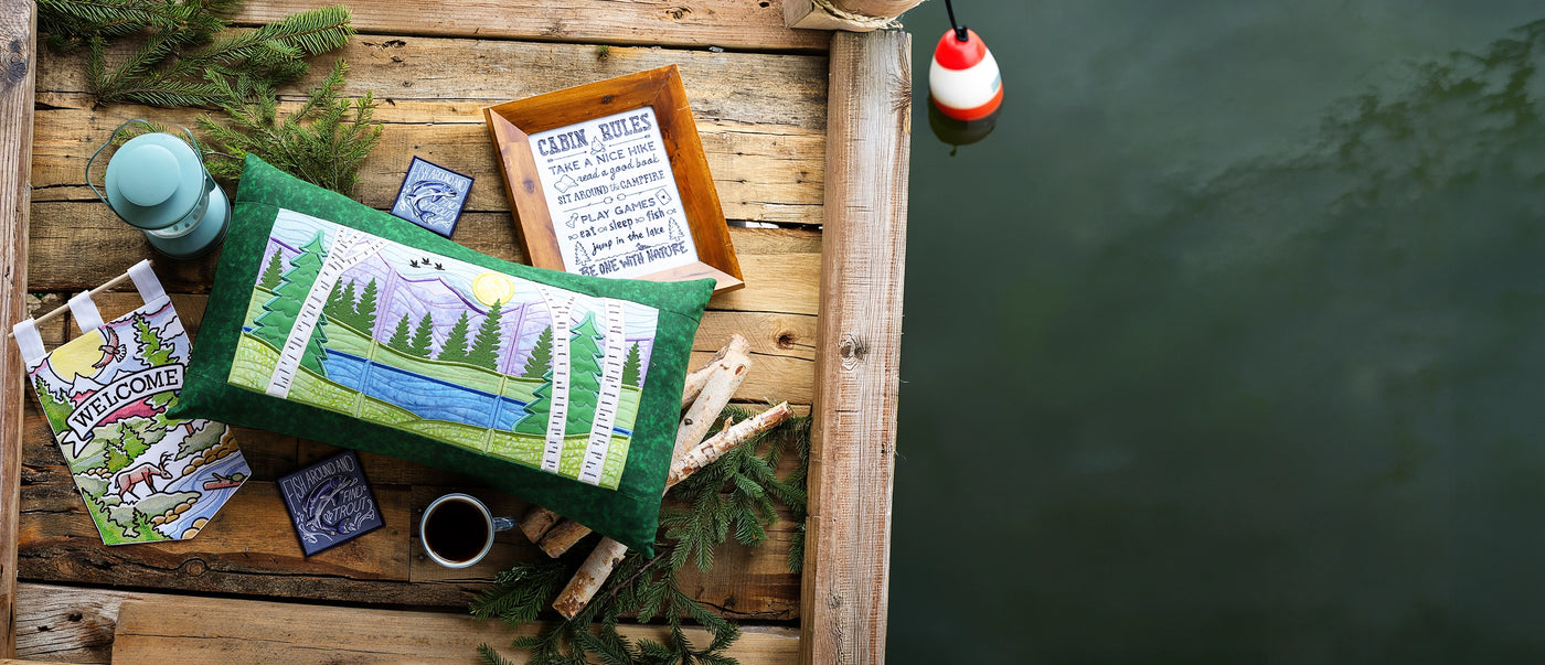 Decorative pillow with nature machine embroidery design staged with other machine embroidery projects on a wooden dock by water