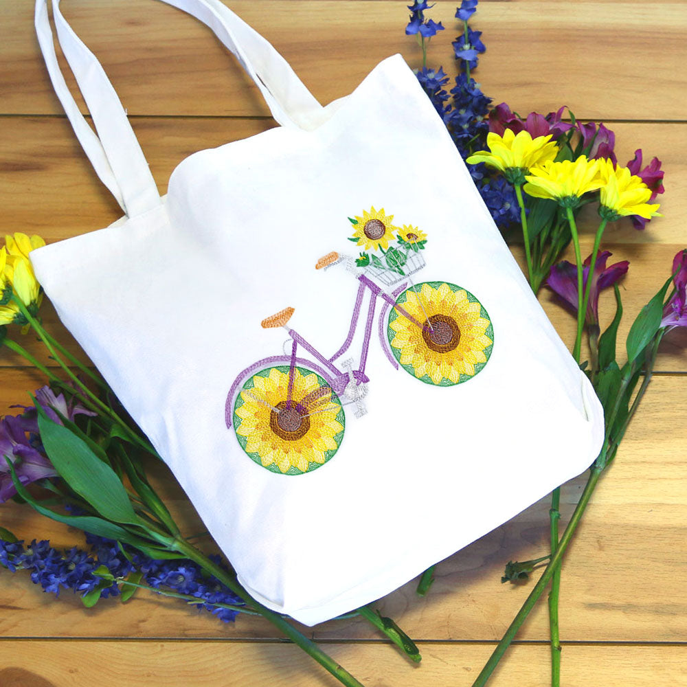 Embroidered bicycle with sunflowers, fruit, and flowers stitched on a white tote bag, surrounded by yellow and purple blooms.