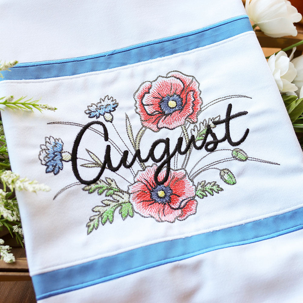 Machine-embroidered red flowers, green leaves, and blue blossoms on white fabric with blue trim, August floral design.