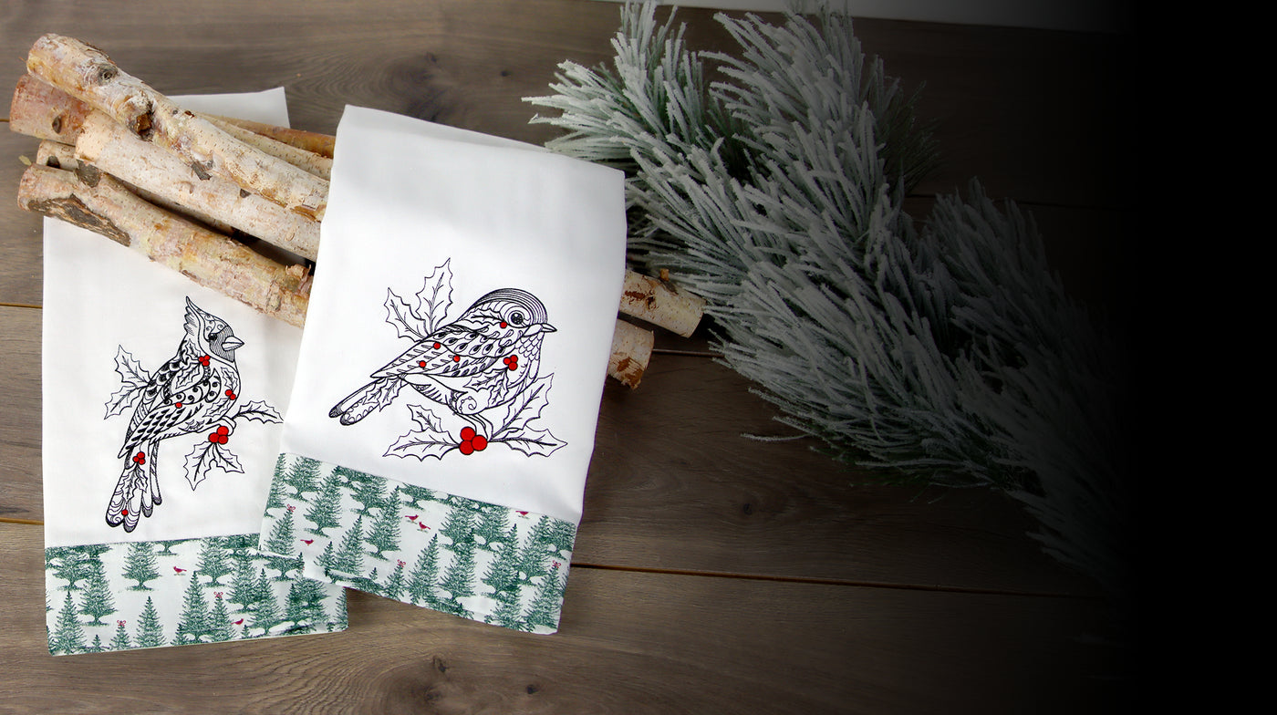 Two machine embroidered towels with cardinal and chickadee designs on a wooden surface with branches and greenery.