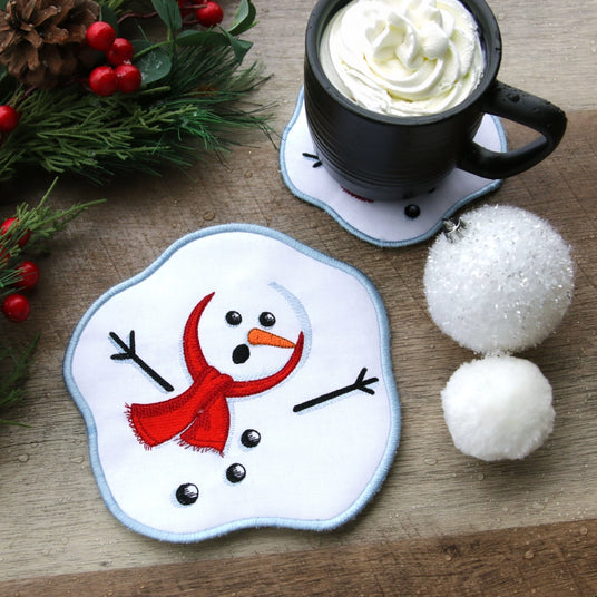 Machine embroidered In-the-Hoop Snowman coaster on a wooden surface with a mug of hot chocolate and decorative items.