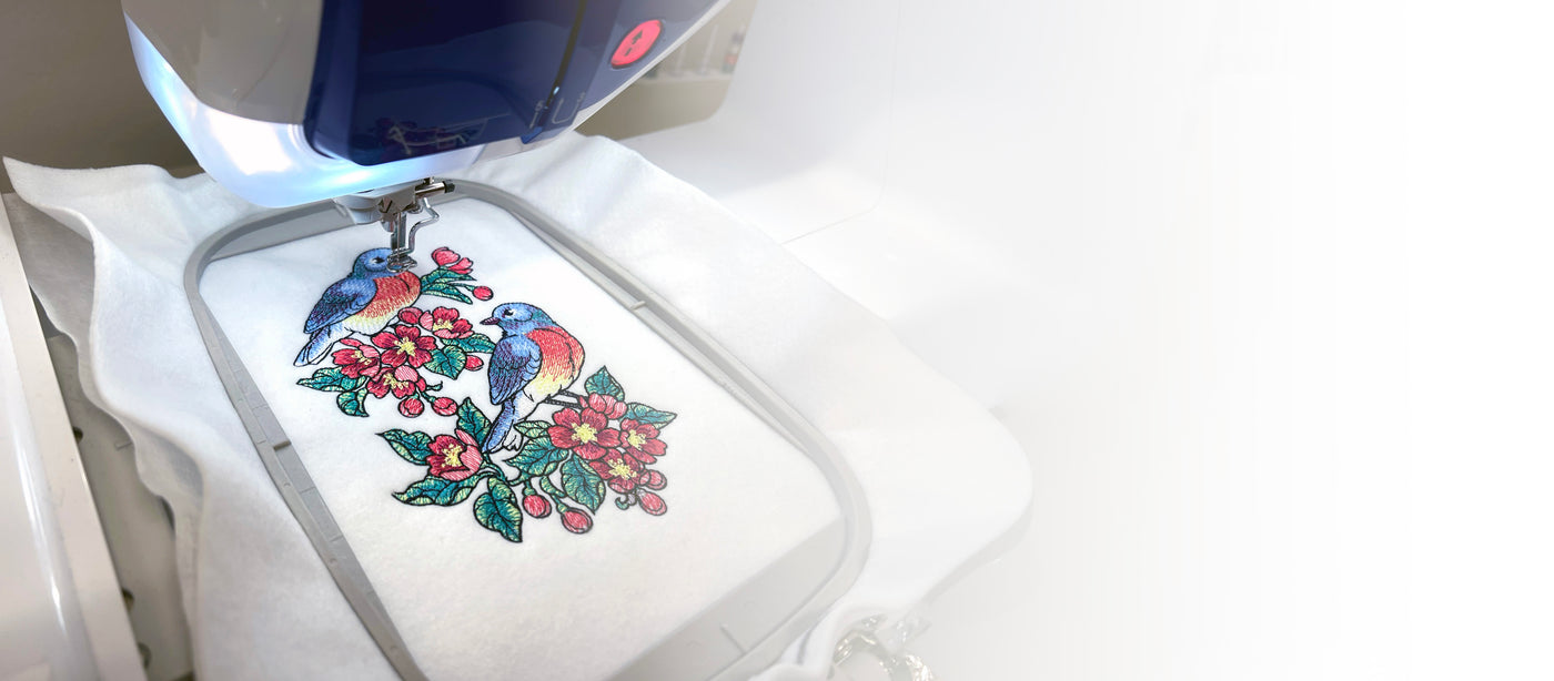 Machine embroidery design of birds and flowers on white fabric under an embroidery machine