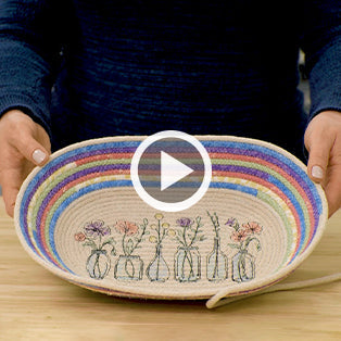 Machine embroidered rope bowl being held up by hands with colorful fabric surrounding rope on rim and wild flowers in vases embroidered on the bottom center - play button on top