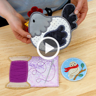 A machine embroidered chicken trivet held by hands over wooden table - a purple thread spool mug rug with a needle and thread and a winter owl coaster design with owl wearing red scarf sitting on branch
