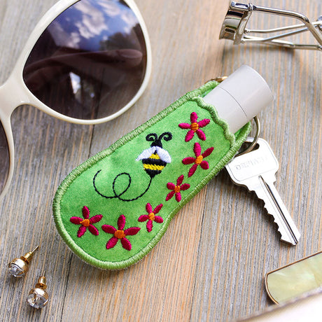 an in-the-hoop machine embroidery design of a keychain lip balm holder featuring a bee and flowers