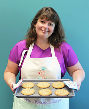 Woman wearing embroidered apron featuring 'Chef Pig' design, and holding a tray of cookies