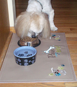 Dog eating from a food bowl sitting on the completed Embroidered Pet Placemat, featuring 'Paws Off!' and 'Top Dog Corner' designs