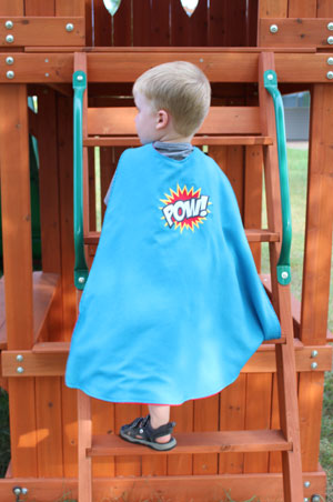Little boy modeling embroidered superhero cape, featuring  'Superhero Pow' design