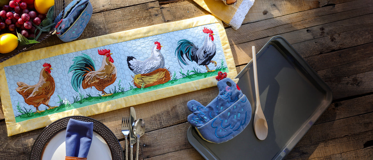 Farmhouse kitchen embroidery projects featuring a quilted table runner with colorful embroidered chickens and roosters, alongside a blue chicken-shaped oven mitt on a rustic wooden table.