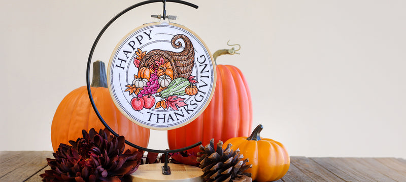 Thanksgiving machine embroidery design featuring a colorful cornucopia filled with pumpkins, fruit, and autumn leaves, surrounded by "Happy Thanksgiving" lettering in an embroidery hoop displayed with fall pumpkins and pinecones.