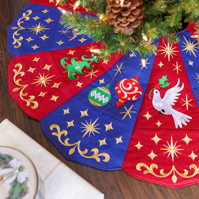 Christmas tree skirt with red and blue panels featuring embroidered gold stars and swirls, green Christmas tree, red and green ornaments, and a white dove holding holly. A decorated pine tree with lights is partially visible above the skirt.