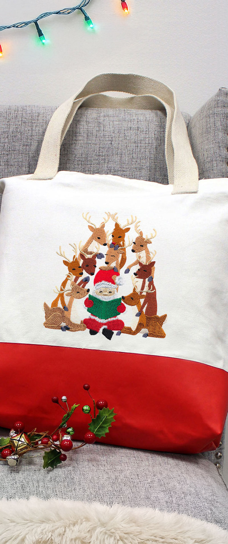 Canvas tote bag with red bottom and beige handles, embroidered with a small Santa reading a green book, surrounded by nine sitting reindeer.