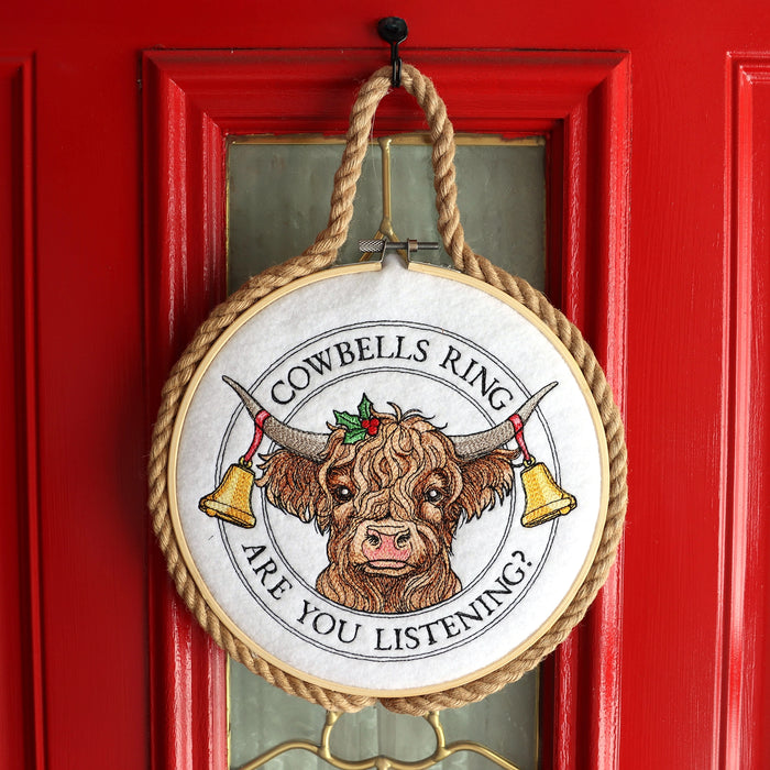 Decorative embroidery hoop sign featuring a machine embroidered highland cow design with text reading 'Cowbells Ring Are You Listening?' on a red door.