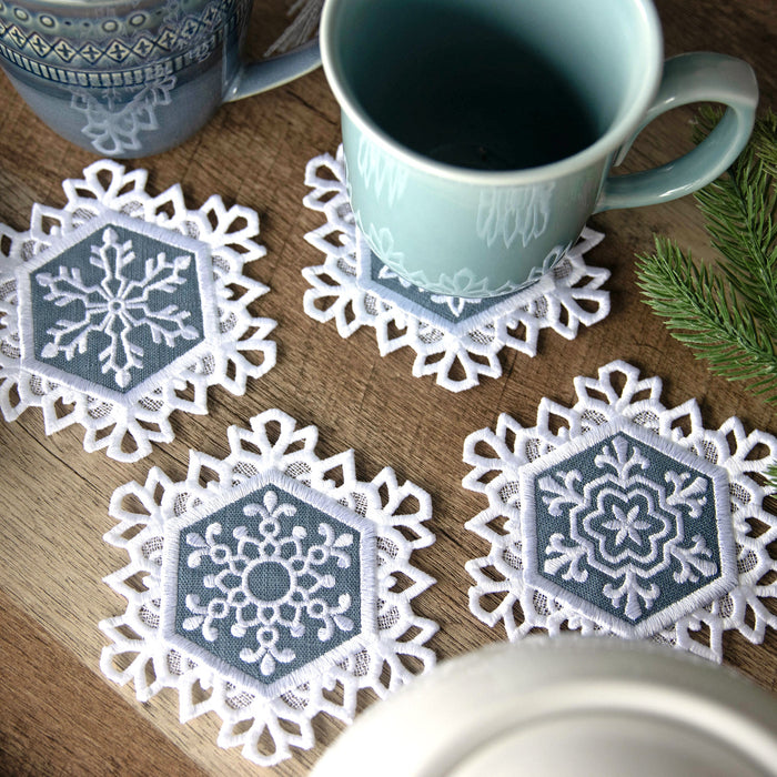 Set of machine embroidery lace edge snowflake coasters on a wooden surface with mugs and greenery.