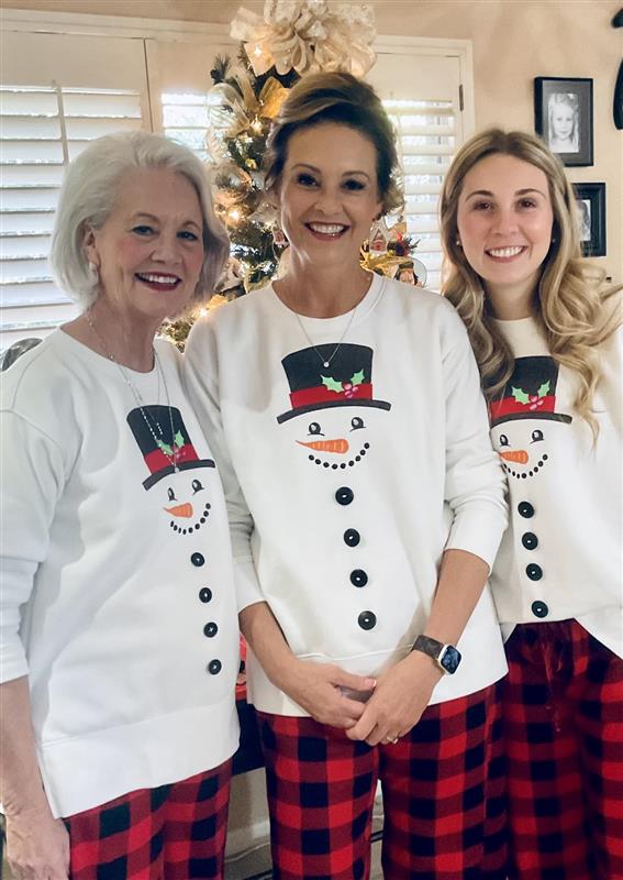 Three women wearing matching machine embroidered snowman design on sweaters and plaid pants in front of a decorated Christmas tree