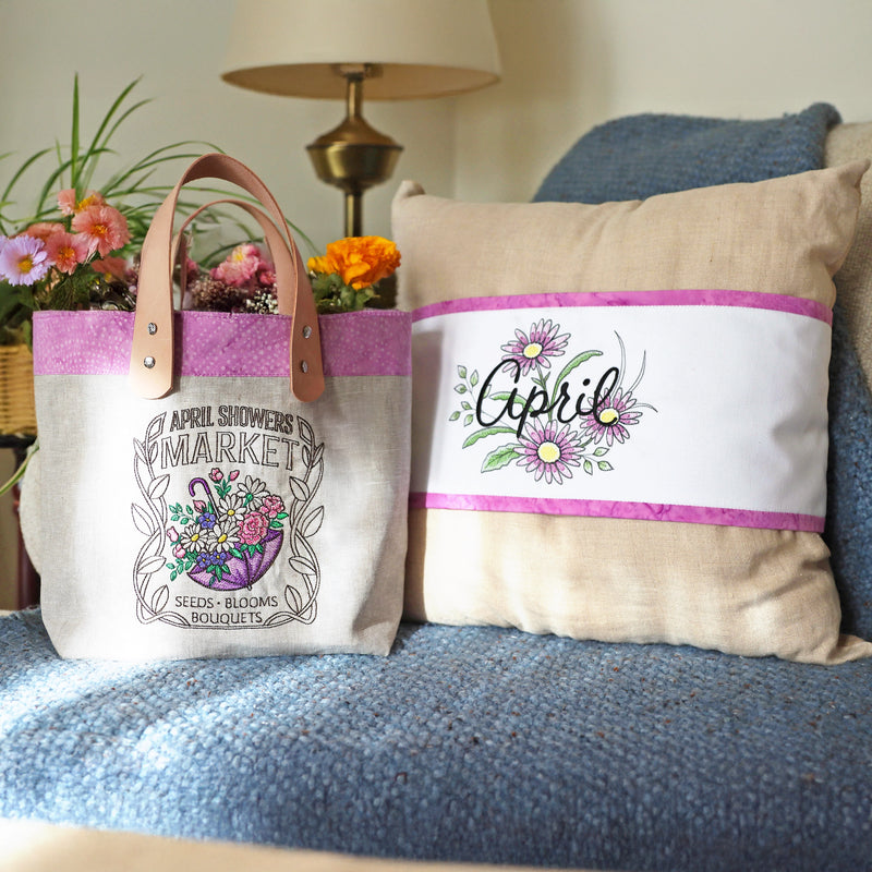 Tote bag and pillow with 'April' inspired machine embroidery designs on a blue couch.
