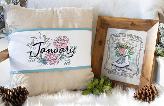 Decorative pillow with machine embroidery 'January' text and floral design, next to a framed machine embroidery frosted winter market design.