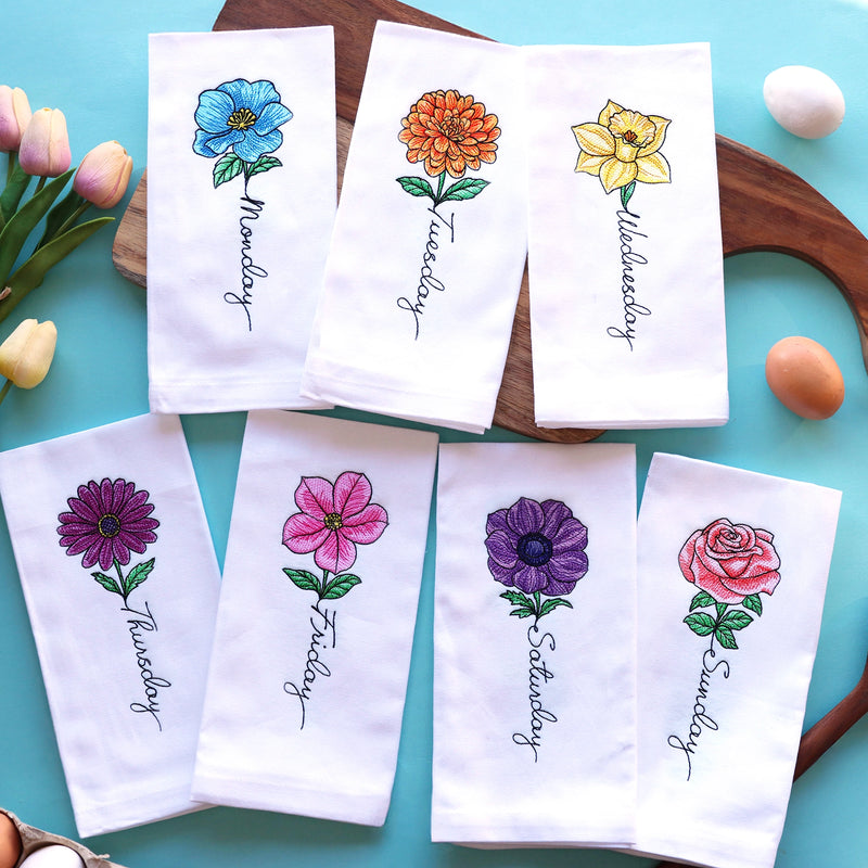 Set of seven white napkins with floral machine embroidery designs and days of the week text on a turquoise surface.