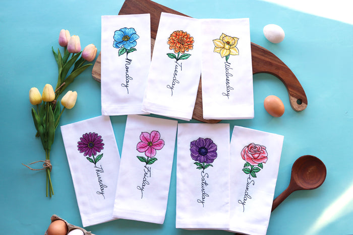 Set of seven white napkins with colorful  machine embroidery floral designs and days of the week text on a blue surface.