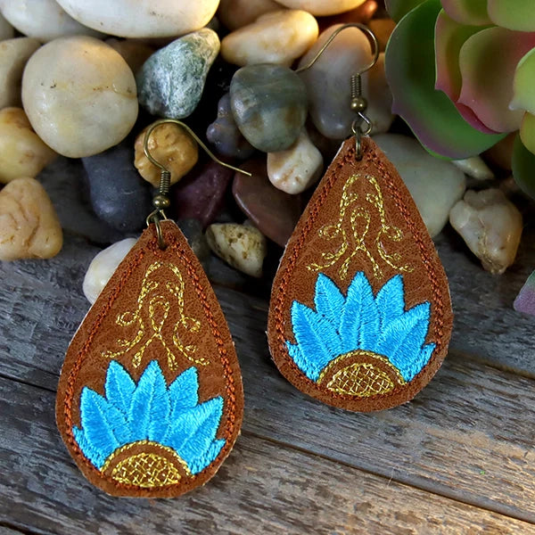Teardrop-shaped  machine embroidered earrings with blue and gold designs on a wooden surface with stones and plants in the background.