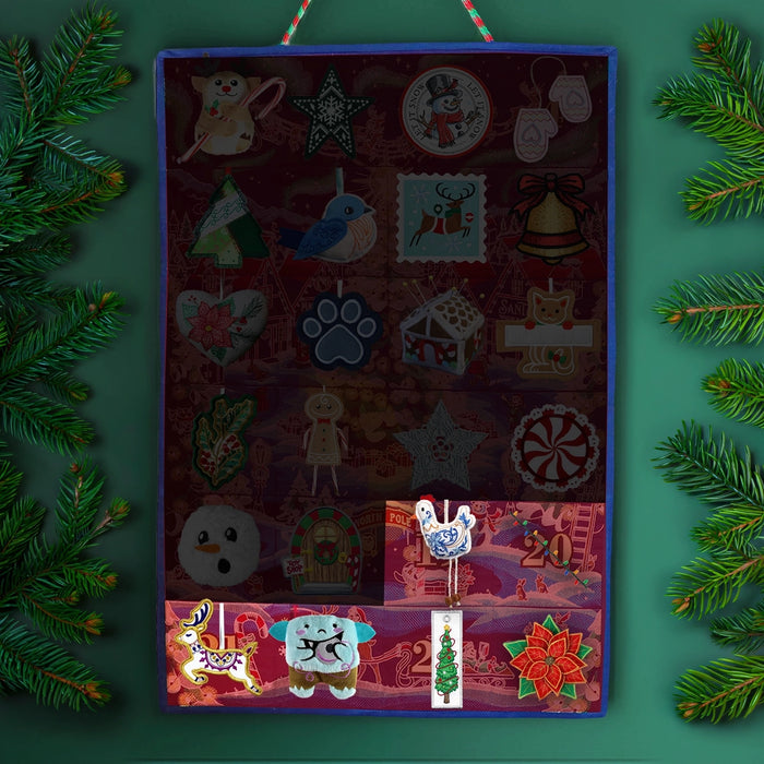A Christmas advent calendar hanging on a green background, decorated with pine branches on the sides. The calendar has multiple colorful pockets and machine embroidered ornaments, including a gingerbread man, a blue bird, a snowman, a Christmas tree, and a red poinsettia.