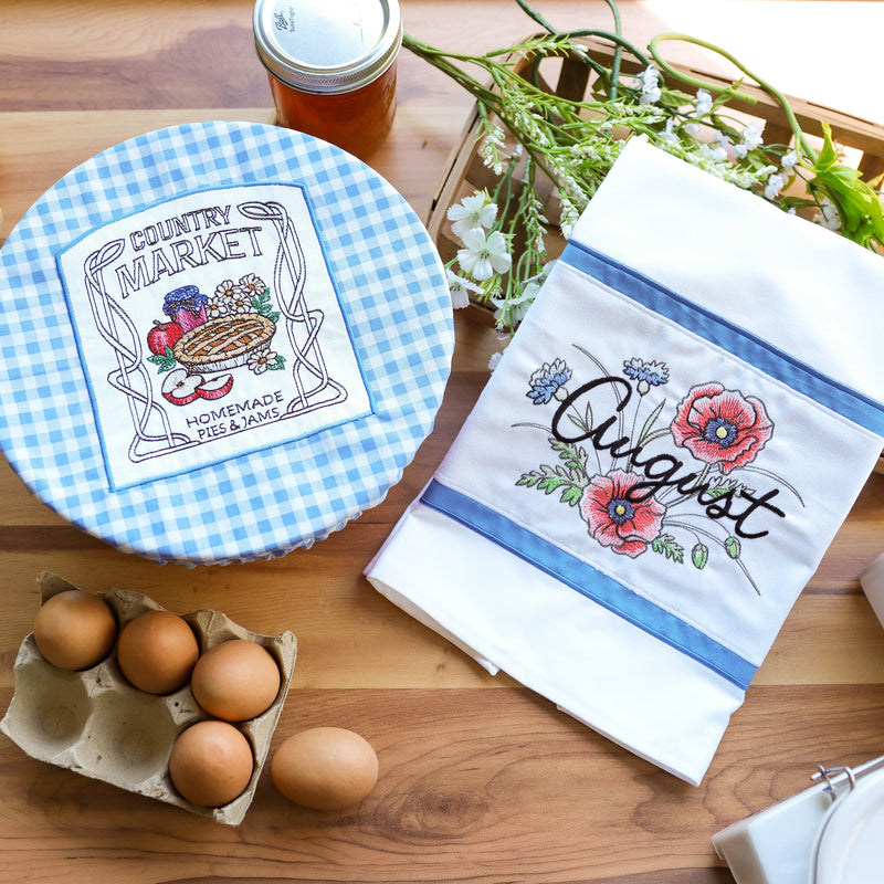 A styled flat-lay of two machine embroidered kitchen textiles on a wooden table: a round blue-and-white gingham covered dish or tray with a central embroidered patch reading 'Country Market Homemade Jam,' and a white tea towel or napkin with blue stripes and floral embroidery reading 'August.' Eggs, a jar, flowers, and a basket are arranged around them as props.