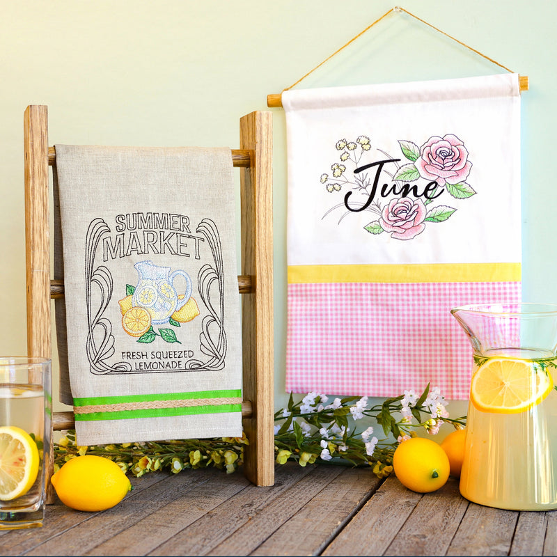 Decorative machine embroidered towel with 'Summer Market' design and a hanging banner with 'June' text, lemons, and flowers on a wooden surface.