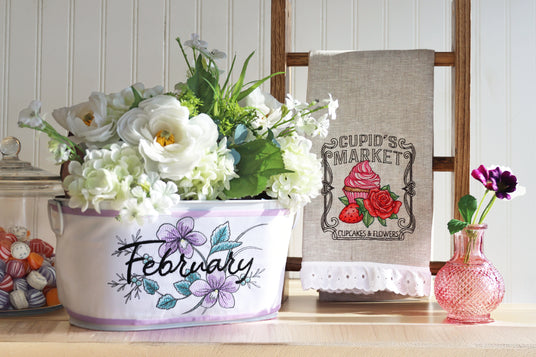 Decorative machine embroidered Cupid's Market design on tea towel next to machine embroidery floral design with 'February' wrapped around a floral basket.