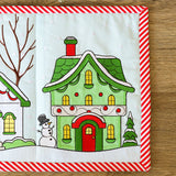 Digitally downloadable machine embroidery design of an evergreen cottage with a snowman, stitched in festive colors.