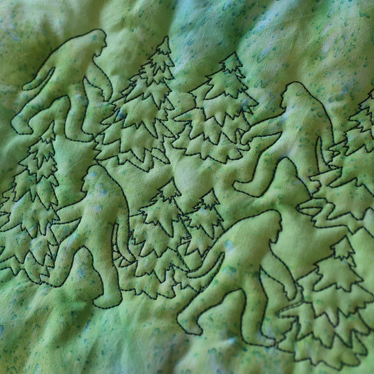 Edge to edge machine embroidery quilting design of sasquatch and pine trees stitched in dark green thread on a light green fabric.