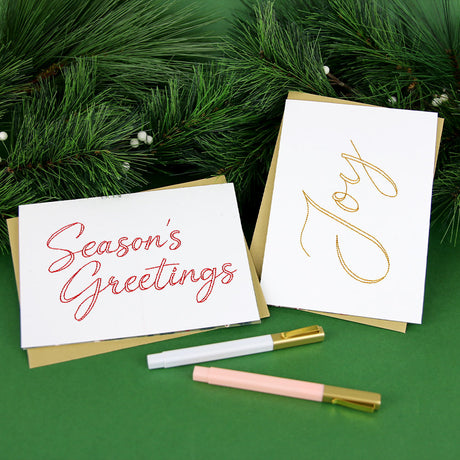 Two white holiday cards with brown envelopes, one embroidered with "Season's Greetings" in red and the other with "Joy" in gold, placed on a green surface with pine branches and two pens nearby.