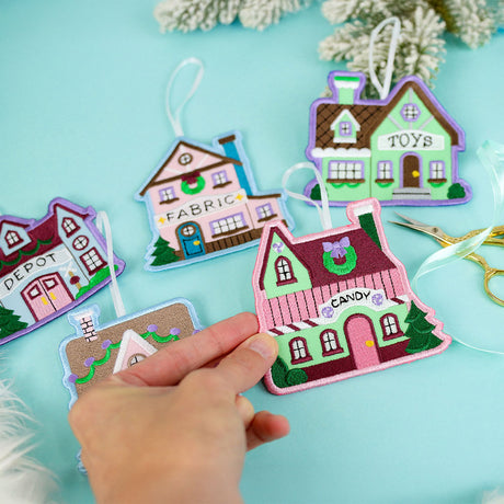 A hand holds a pink and green embroidered house ornament labeled "CANDY" with other house ornaments labeled "FABRIC," "DEPOT," and "TOYS" on a pale blue background.