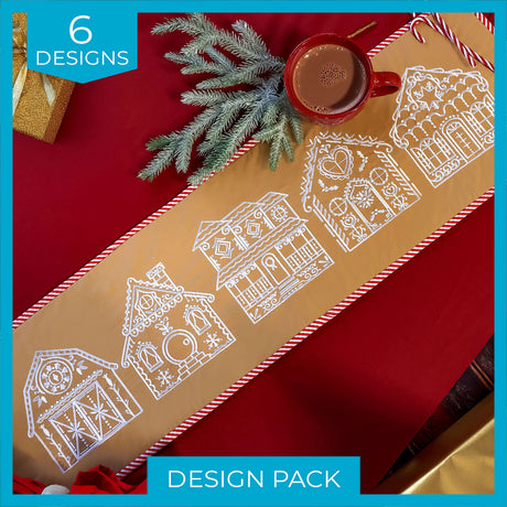 A beige table runner with white embroidery of five gingerbread house designs on a red background with a red mug of hot chocolate and evergreen sprigs.