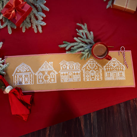 Table runner with white embroidered gingerbread-style houses on a tan background, bordered with red and white stripes, placed on a red cloth with pine branches and a red cup of cocoa.