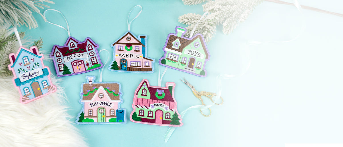 Six colorful embroidered Christmas village house ornaments labeled Bakery, Depot, Fabric, Toys, Post Office, and Candy, laid on blue background with white branches.