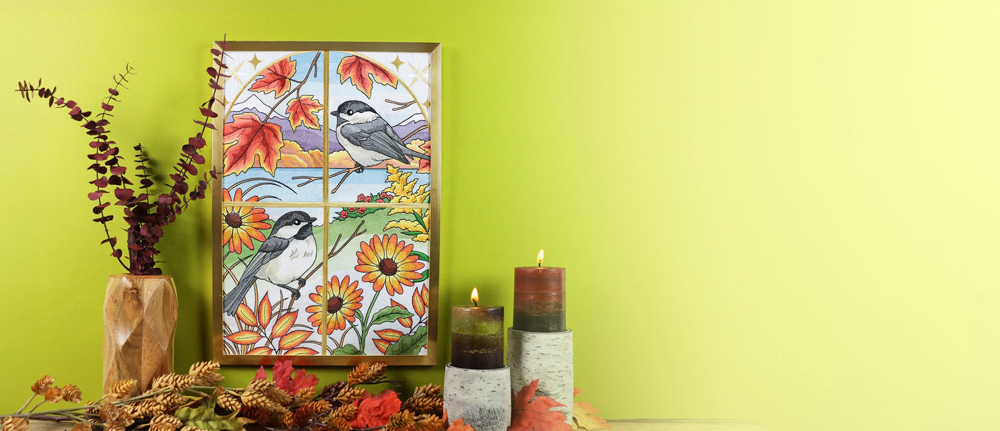 A framed autumn-themed embroidery scene featuring two black-capped chickadees and colorful fall leaves and sunflowers, displayed on a light green wall alongside candles, a vase with dried stems, and autumn decorations.
