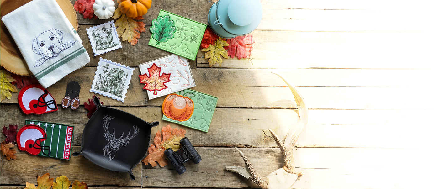 A styled flat-lay of multiple autumn-themed machine embroidery projects and finished items arranged on a rustic wooden surface with fall leaves, pumpkins, and antlers. Visible items include patterned fabric squares, a white towel with a dog design, a black zip pouch with a deer head print, small fabric accessories, and seasonal decorations.