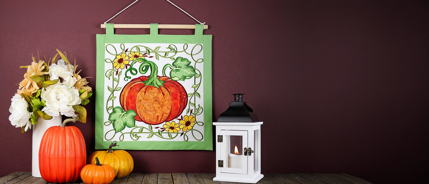 A decorative hanging fabric wall tapestry with a machine embroidered pumpkin design, displayed on a wooden surface next to a white vase of flowers, small pumpkins, and a white lantern against a dark burgundy wall.
